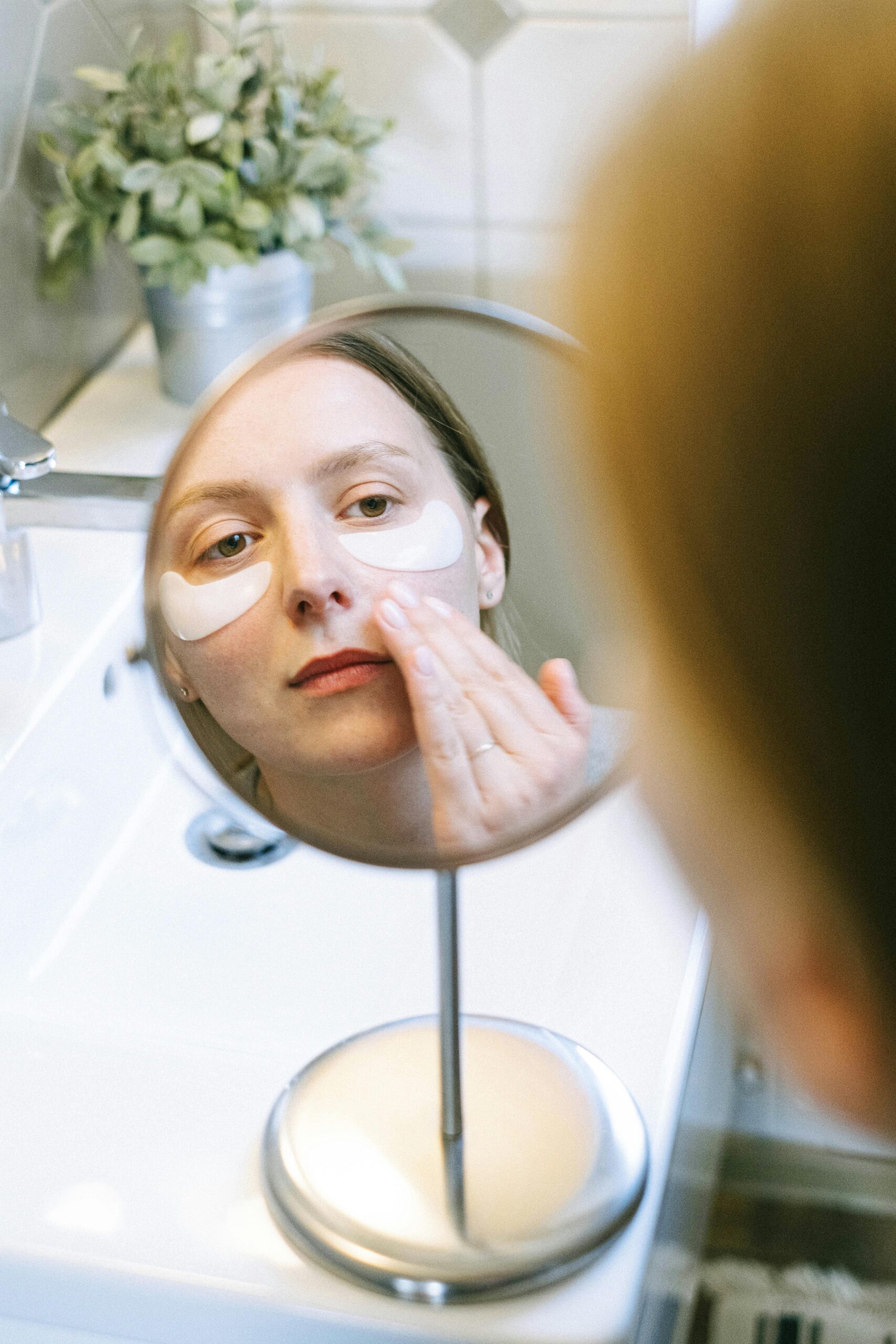 Woman applying eye patches as part of her skincare routine, reflected in bathroom mirror.